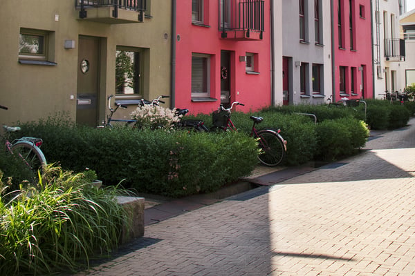 Bild på en radhuslänga där fasaden skiftar i beige, rött och vitt. Framför står några cyklar parkerade mellan gröna buskar.